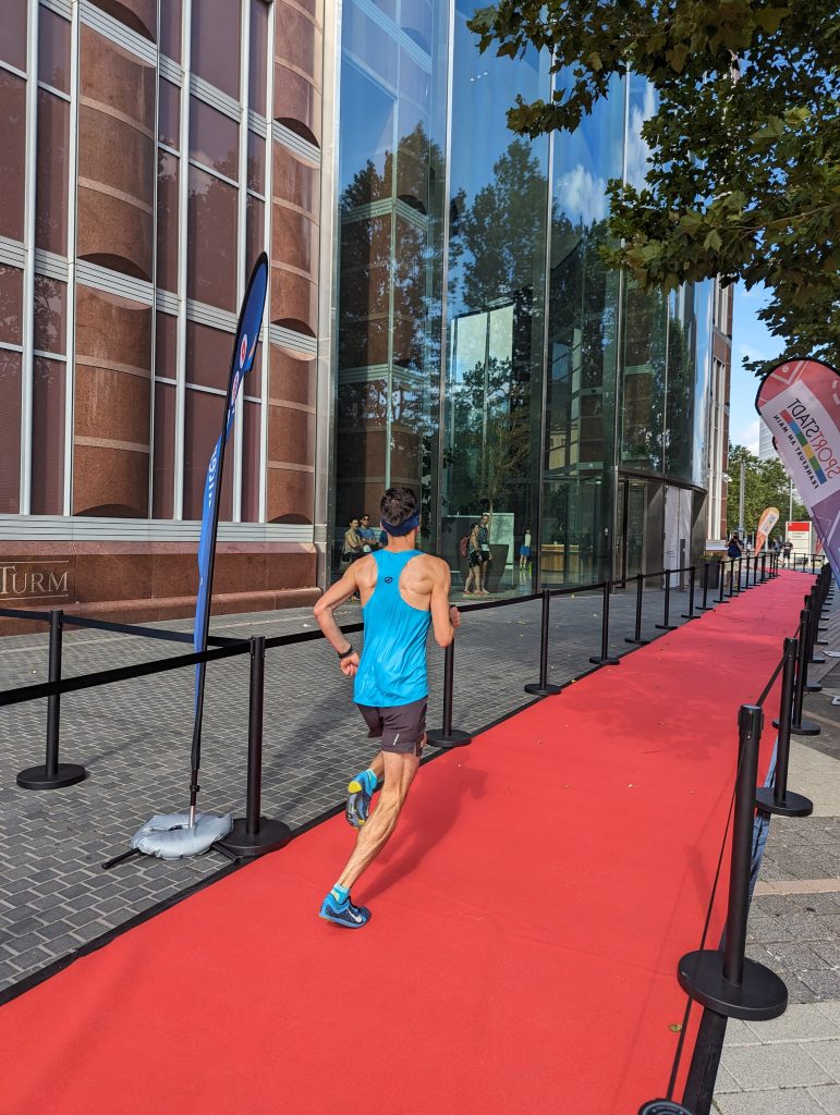Ein Läufer in blauem Trikot von hinten auf dem blauen Teppich vor dem Haupteingang des Messeturms